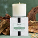 A lit white candle in a clear glass vessel sits on top of its white box packaging, which reads 'LAFCO NEW YORK Feu de Bois'. The scene is styled with rustic driftwood, burning wood, and small stones against a textured green background with smoke rising.