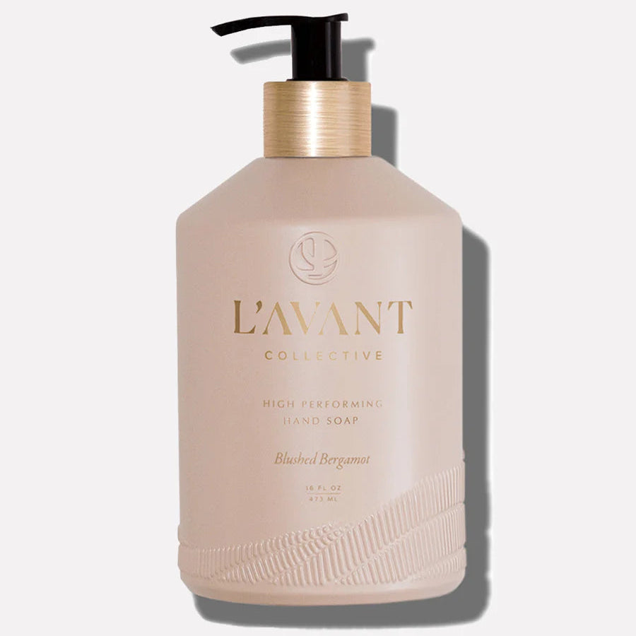 A pink glass bottle with a gold pump containing hand soap, labeled 'L'AVANT Collective', with a hint of shadow on the bottom right.